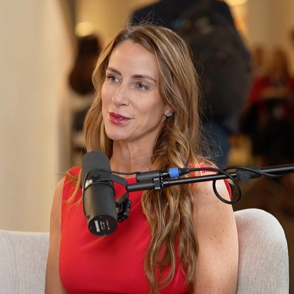 A woman with long, wavy, red hair talking into a microphone during an interview or podcast recording in a cozy indoor setting.